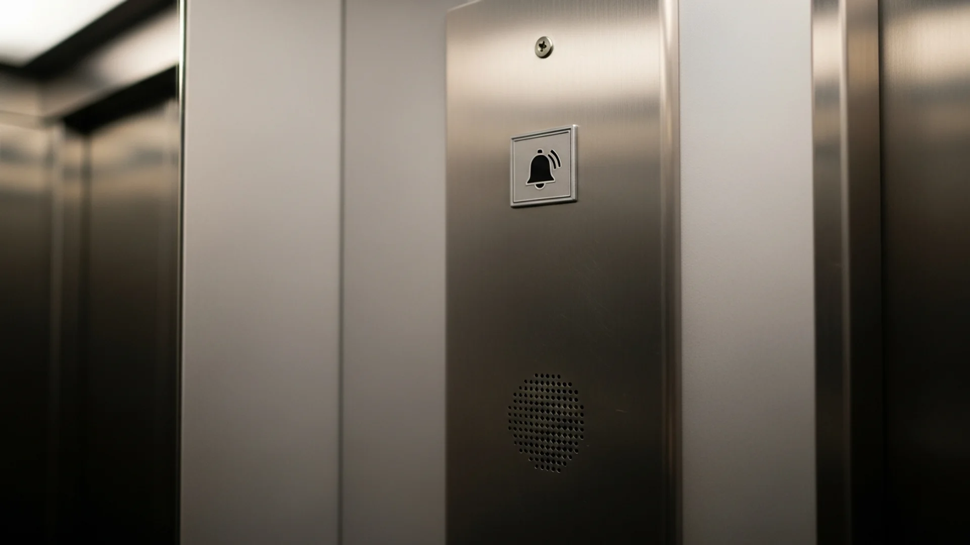 Close view of the stainless-steel control panel inside a passenger lift car, showing a raised emergency call button marked with a bell symbol and a speaker grille